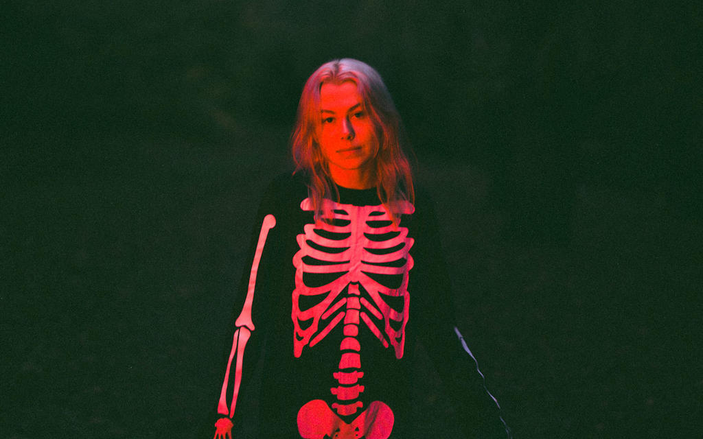 Phoebe Bridgers, seen wearing her iconic skeleton suit, bathed in glowing red light in the midst of night.