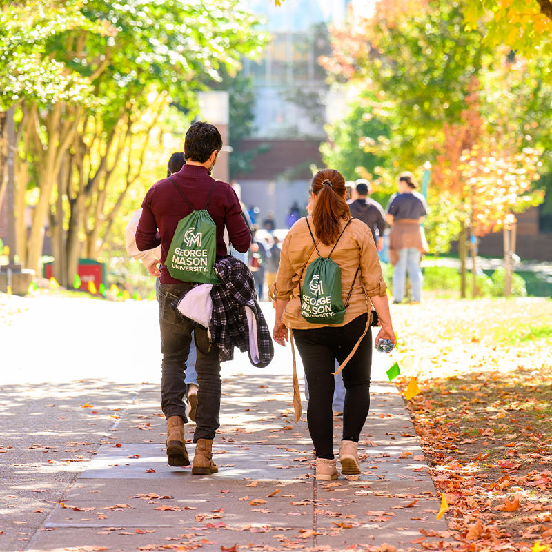 A prospective student and their family member walk down a sunlit pathway on the Fairfax Campus, flanked on both sides by fall foliage.